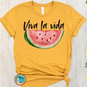 Peut inclure: Un t-shirt jaune avec le texte "Viva la vida" et un graphique de tranche de pastèque. La tranche de pastèque a des pépins noirs et le texte "Frida Kahlo" est écrit en bas.