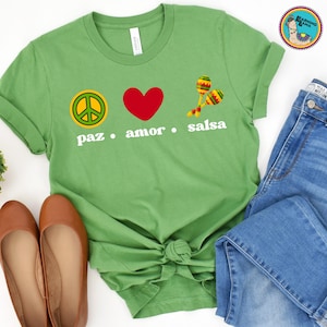 May include: A green t-shirt with the words "paz, amor, salsa" and peace sign, heart, and maracas graphics. The shirt is paired with blue jeans and brown flats. A round logo is in the upper right corner.