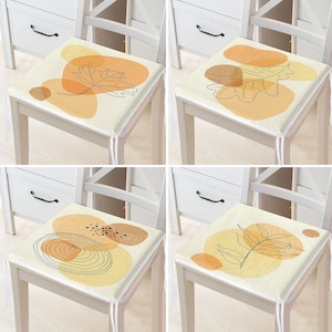 May include: Four square chair cushions with abstract leaf designs. Each cushion features a cream background with orange and yellow shapes and a black outline of a leaf. The cushions are tied to white chairs.