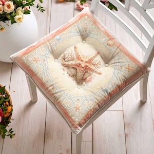 May include: A square chair cushion featuring a central starfish and seashell illustration on a cream background. The design includes a light blue rope border, intricate scrollwork, and a soft peach-colored frame with tufted button accents.