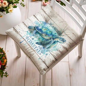 May include: A square chair cushion featuring a watercolor sea turtle illustration on a white wood-grain background. The design includes blue paint splatters, small starfish, and seashell motifs with the text WANDER and COASTAL printed in blue.