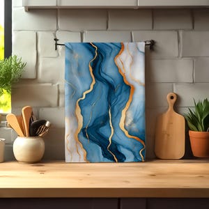 May include: A rectangular kitchen towel featuring a fluid, abstract pattern of deep blue, light blue, and white swirls accented with thin, metallic gold veins. The towel hangs from a black metal bar against a white brick-style kitchen backsplash.