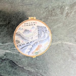 May include: A round, hinged trinket box with a gold-colored rim. The lid features a painted scene of the Great Wall of China, with text in Chinese characters. The background is a marble surface.