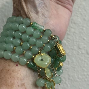 May include: A multi-strand bracelet featuring smooth, light green jade-like beads interspersed with small gold-toned metal spacers. The bracelet includes dangling charms, including a gold-toned peanut shape and carved light green stone pendants.