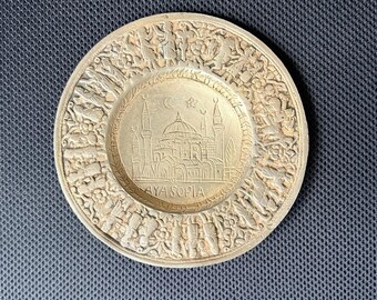 May include: A round, silver-colored decorative plate with a detailed relief design. The center features a building with minarets, a crescent moon, and a star. The word "AYASOFIA" is inscribed below the building. The outer rim has a detailed relief of figures and patterns.