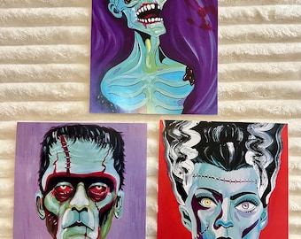 May include: Three Halloween-themed art prints featuring classic monster portraits. The top print depicts a zombie with a teal complexion against a purple backdrop. The other two prints show Frankenstein's monster and his bride.