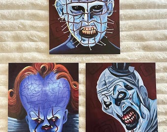 May include: Three horror-themed art prints. The top print features a blue-toned portrait of a character with nails in their head. The bottom left print is a blue clown portrait with red hair. The bottom right print is a white and blue clown portrait.