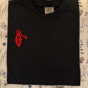 May include: A black short-sleeved t-shirt featuring a red embroidered graphic on the left chest. The graphic depicts a vintage-style hotel key tag with an infinity symbol and a key. The inner neck label displays the text Comfort Colors.