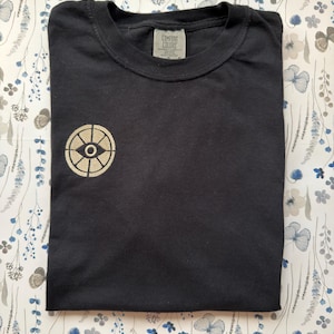 May include: A black short-sleeve t-shirt featuring a circular beige embroidered emblem on the left chest. The emblem depicts a stylized eye within a segmented wheel design. The shirt has a crew neckline and a Comfort Colors brand label.