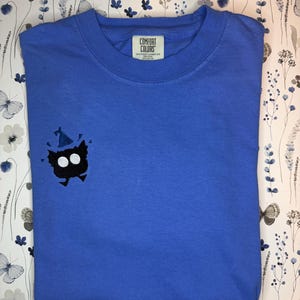Op de afbeelding: Een blauw t-shirt met ronde hals en een kleine, zwart geborduurde creatuur met twee grote witte ronde ogen en een blauwe feesthoed op het hoofd. Het shirt heeft een Comfort Colors-merklabel aan de binnenkant van de kraag, wat 100% katoen aangeeft.