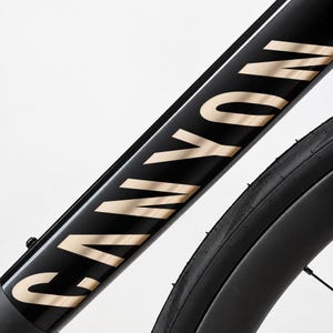 Op de afbeelding: Een zwart fietsframe met de merknaam CANYON in grote, metallic gouden hoofdletters langs de diagonale buis. Een deel van een zwarte band met een gestructureerd profiel is zichtbaar aan de rechterkant van het frame.