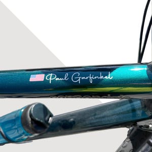 Op de afbeelding: Een donkergroen-blauwe metallic fietsframe met een witte sticker waarop de naam Paul Garfinkel in schrijfletter staat, naast een klein Amerikaans vlaggetje. Het frame heeft een glanzende afwerking en een bijpassende bidon is gedeeltelijk zichtbaar.