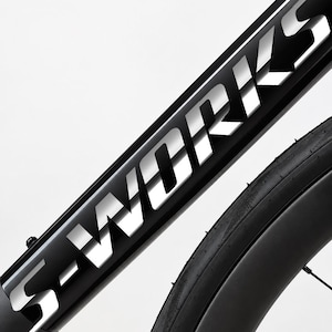 Könnte beinhalten: Ein schwarzer Fahrradrahmen mit dem Schriftzug S-WORKS in großen, reflektierenden silbernen Großbuchstaben entlang des diagonalen Rohrs, neben einem schwarzen Gummireifen mit sichtbarem Profil und dünnen schwarzen Speichen.