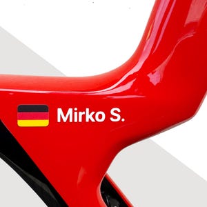 May include: A glossy red bicycle frame section featuring a small rectangular German flag icon next to the white text Mirko S. The frame has a smooth, reflective surface with a black accent visible on the left side of the curved structure.