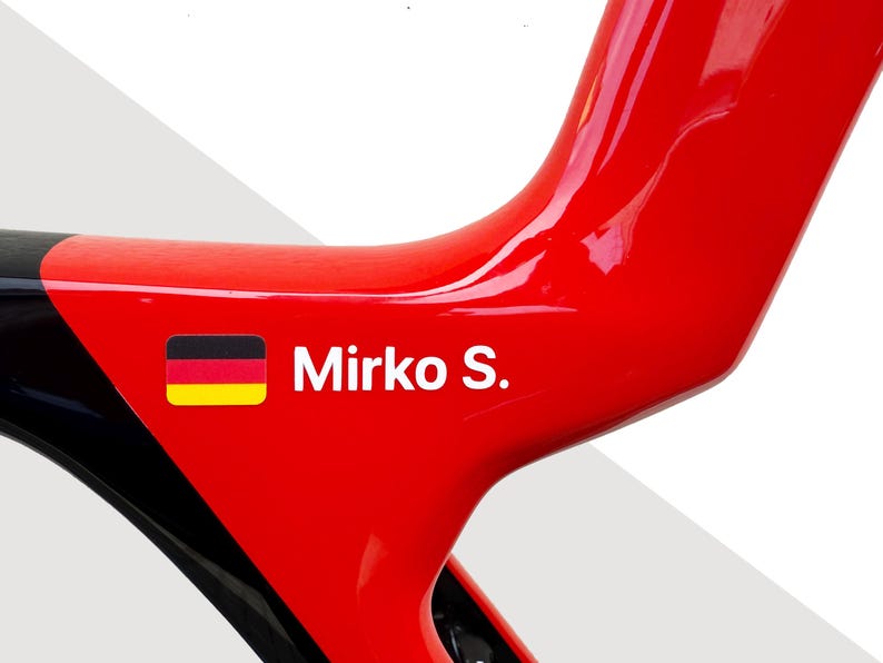 May include: A glossy red bicycle frame section featuring a small rectangular German flag icon next to the white text Mirko S. The frame has a smooth, reflective surface with a black accent visible on the left side of the curved structure.