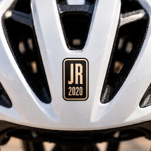 May include: A white bicycle helmet with black interior ventilation channels and a rectangular black sticker on the front centre. The sticker features a gold border and the gold text JR 2020 stacked vertically in a clean, sans-serif typeface.