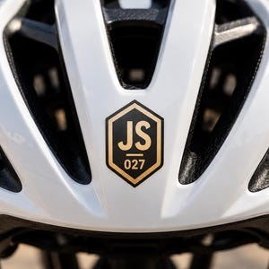 May include: A white bicycle helmet with multiple ventilation slots and a black interior lining. Centered on the front is a black hexagonal emblem with a gold border, featuring the letters JS above the number 027 in a gold sans-serif font.