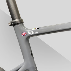 May include: A close-up of a grey bicycle frame with a black seat post. The frame has the text "Kevin MORRIS" with a Union Jack flag. The bike appears to be a road bike.