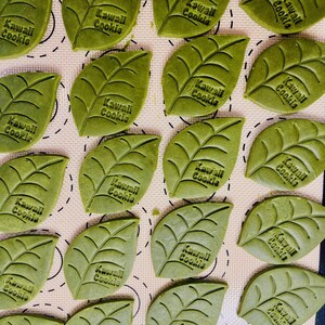 May include: A baking sheet filled with green leaf-shaped cookies, each decorated with the text "Kawaii Cookie".