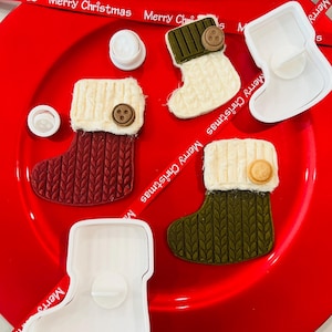 May include: Three Christmas stocking cookie cutters, two white and one green, with a red, white, and green Christmas stocking cookie on a red plate. The cookie cutters are made of plastic and have a smooth finish. The cookies are decorated with white icing and a brown button.