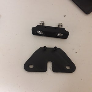 May include: Two black 3D printed plastic parts for a 3D printer. The top part has two silver screws and two silver nuts. The bottom part has a U-shaped opening and two silver screws.