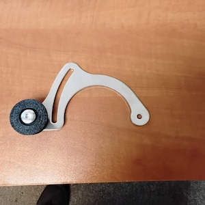 Può includere: Un componente metallico color argento con un braccio curvo e una piccola ruota nera e rotonda attaccata. La ruota ha un centro argentato. Il componente è posto su una superficie di legno marrone chiaro.