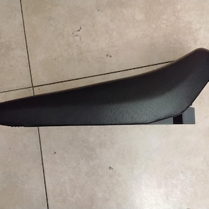 May include: A black motorcycle seat with a textured surface. The seat has a curved design and is elevated on a small, dark-coloured support. The seat is designed for comfort and support during rides.