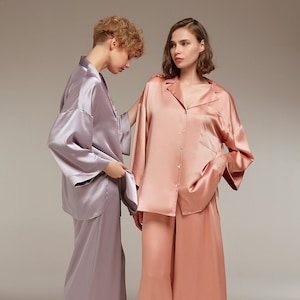 May include: Two women wearing matching silk pyjamas. The woman on the left is wearing a lavender set, and the woman on the right is wearing a peach set. Both sets feature a collared shirt with a button-up front and wide-leg trousers.