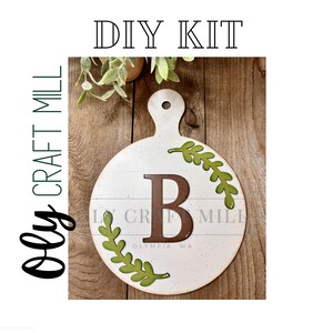 May include: DIY kit for a round wooden cutting board with a white background. The board has a brown letter "B" in the center surrounded by green leaves. The text "DIY KIT" is at the top of the image and "Oly Craft Mill" is at the bottom left.