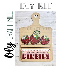 May include: DIY craft kit featuring a wooden cutting board with a heart-shaped handle. The board has three strawberry cutouts and a sign that reads "barn fresh berries".