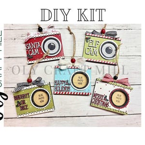 May include: DIY Christmas camera ornaments with festive text: "Santa Cam," "Elf Cam," "Happy Holidays," "Naughty or Nice," and "Merry Christmas." Each ornament has a space for a photo and is decorated with ribbons.