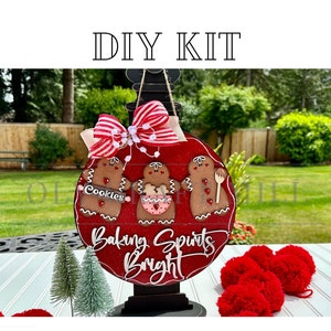May include: DIY kit for a Christmas ornament featuring three gingerbread people with the text "Baking Spirits Bright" on a red background. The ornament is on a black stand and there are red pom-poms in the foreground.