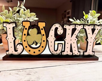May include: Wooden sign with the word "LUCKY" in pink and white polka dot letters. The letter "U" is a horseshoe with a green shamrock inside. The sign is on a wooden base.