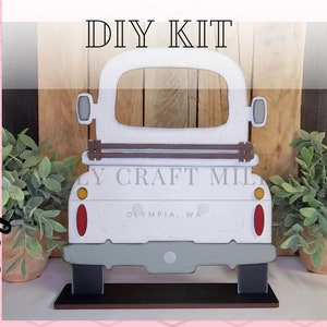 May include: DIY kit for a white wooden truck cutout with a brown tailgate and a gray bumper. The truck has a window cutout and the text "OLY CRAFT MILL OLYMPIA, WA" is printed on the back.