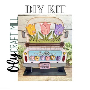 May include: DIY craft kit featuring a wooden truck with a white cab and a blue bed. The truck is decorated with three colorful tulips and the words "hello spring" and "flower market".