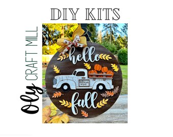 May include: A DIY craft kit featuring a wooden round sign with the words "hello fall" and a white truck carrying pumpkins. The sign is decorated with fall leaves and a yellow and red bow.