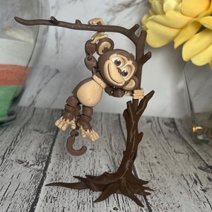 May include: A brown monkey figurine hanging from a brown tree branch. The monkey has a smiling face and is holding a banana. The tree has a brown base.