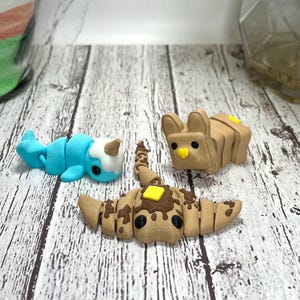 May include: Three 3D-printed toys: a blue narwhal with a white horn, a brown croissant with chocolate and butter, and a brown bunny with a yellow square. The toys are on a light wood-grain surface.