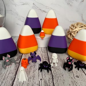 May include: A set of six candy corn shaped Halloween decorations, each with a white top, orange middle, and purple bottom. The decorations are arranged in a circle around a small white ghost figurine. There are also several small Halloween figurines, including a bat, a spider, a skeleton, and a pumpkin.