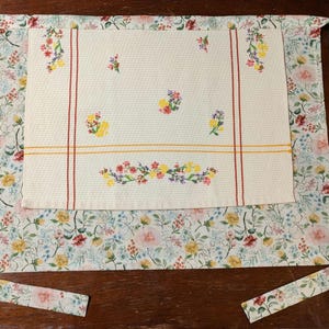 May include: A floral apron with a cream-colored center panel featuring embroidered flowers and red and yellow stripes. The apron's main body has a floral pattern with pink, yellow, and blue flowers. The apron has long ties.