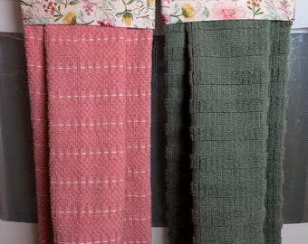 May include: Two hanging kitchen towels featuring a floral print top section with pink, yellow, and green botanical patterns. One towel is a solid dusty pink, and the other is a solid sage green. Both towels have a textured, waffle-like weave.