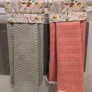 May include: Two kitchen towels hanging on a white appliance. One is sage green with a textured pattern, and the other is coral with a woven design and white lace trim. The top of each towel features a floral pattern on a cream background.