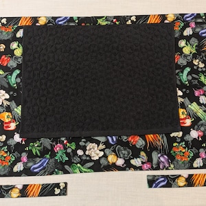 May include: A rectangular waist apron featuring a black background with a colorful vegetable print including carrots, eggplants, peppers, and mushrooms. A solid black quilted fabric panel is centered on the front, and the apron has long black fabric ties.