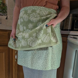 May include: A sage green kitchen towel apron featuring a white pumpkin and leaf pattern on the top section and a white polka dot pattern on the bottom section. The towel is folded over at the waist, showcasing the two distinct fabric designs.