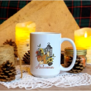 May include: White ceramic mug with a fall-themed design featuring pumpkins, sunflowers, and a lantern. The text "Autumn" is printed in orange on the mug.