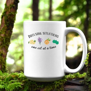 May include: White ceramic mug with a black graphic that says "Diffusing Situations one oil at a time". The graphic also includes illustrations of a lemon, lavender, orange, and a green leaf.