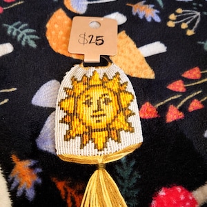 May include: A handmade, off-white and yellow sun-shaped woven keychain with a smiling face. The sun design is in shades of yellow and brown, with a yellow tassel at the bottom. The keychain is displayed against a dark background.