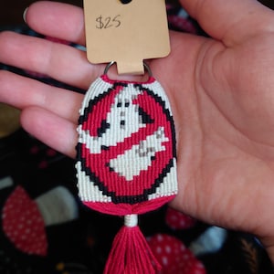 May include: A handmade keychain featuring the Ghostbusters logo in red, black, and white. The keychain has a red tassel and a metal ring for attaching keys. The tag reads $25.