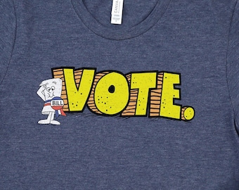 Pode incluir: Uma t-shirt de manga curta em azul marinho mesclado com o texto amarelo VOTE e uma personagem de desenho animado de um documento com uma faixa vermelha, branca e azul. A t-shirt é apresentada com ténis pretos e calças de ganga azuis.