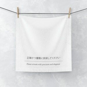 Puede incluir: Toalla de algodón blanca colgada de un tendedero con el texto "正確かつ優雅に排尿してください" en japonés y "Please urinate with precision and elegance" en inglés.